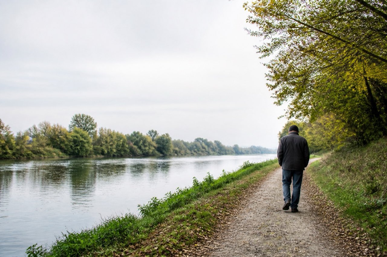 A person in a dark sweater walking beside a riverside path, photographed from behind with room for quiet reflection