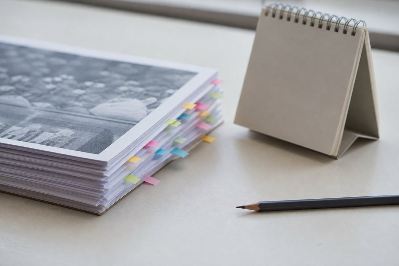 A kitchen table in morning light, an upper-arm blood pressure cuff, a paper log with handwritten dates and two columns of numbers, and a glass of water, shot from slightly above.